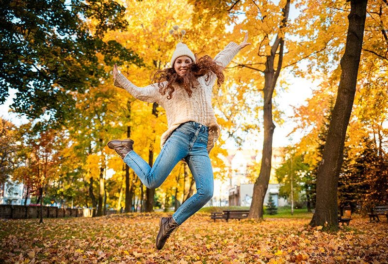 Glückliche junge Frau springt mit erhobenen Armen auf bunten Herbstblättern