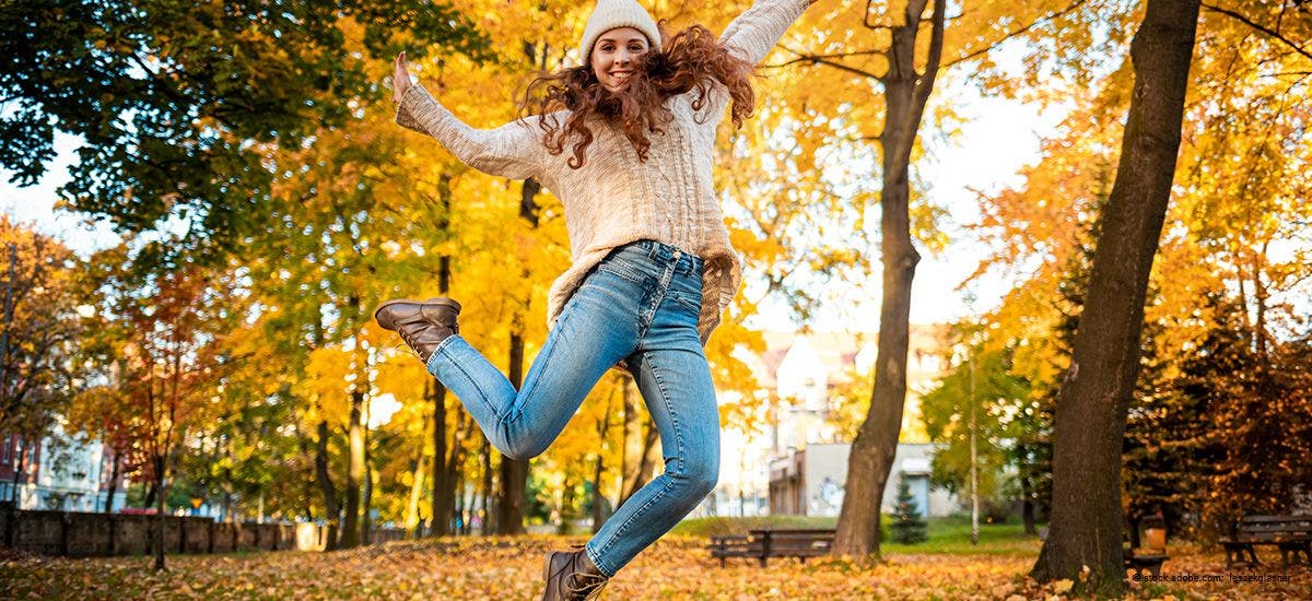 Glückliche junge Frau springt mit erhobenen Armen auf bunten Herbstblättern
